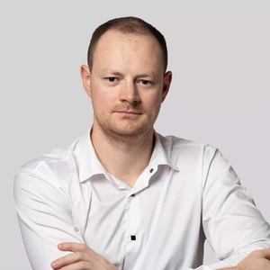 Portrait photo of a man with short hair, wearing a white shirt. He is looking at the camera with a serious expression, arms crossed over his chest, against a neutral grey background.