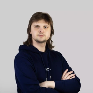 Portrait photo of a young man with medium-length hair, wearing a dark blue hoodie. He is standing with arms crossed, looking at the camera with a serious expression. The background is neutral light grey.