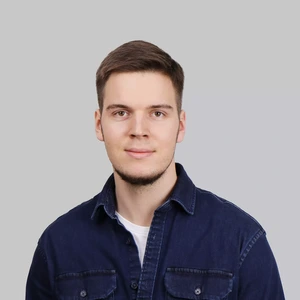 Portrait photo of a young man with short hair, wearing a denim jacket and a white T-shirt. He is looking at the camera with a slight smile, against a neutral grey background.