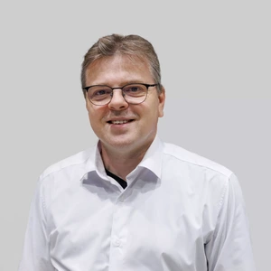 A man with glasses and short hair, wearing a white shirt, looking at the camera and smiling.