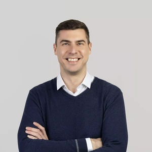 A man with short dark hair, wearing a white shirt and a dark blue sweater, stands with arms crossed and smiles broadly while looking at the camera against a light background.