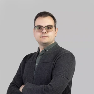 Portrait photo of a young man with short hair and glasses, wearing a dark sweater and a green shirt, looking at the camera with arms crossed against a light background.