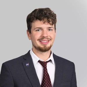 Portrait photo of a young man with curly hair and a beard, wearing a dark blazer, white shirt, and a burgundy polka-dot tie, smiling at the camera against a light background. A NASA pin is attached to the lapel of his blazer.