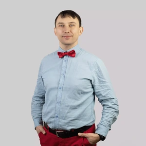 Portrait photo of a man with short dark hair, wearing a light blue shirt, a bright red bow tie, and red trousers. His hands are in his pockets, he is looking straight at the camera. The background is neutral light grey.