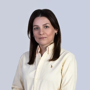 Portrait photo of a young woman with dark hair, wearing light yellow striped shirt, looking at the camera against a light background. She is wearing hoop earrings and has a subtle smile.