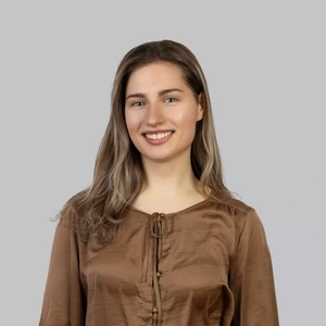 Portrait photo of a young woman with long light brown hair, wearing a brown blouse with a front tie detail. She is smiling while looking at the camera, against a neutral grey background.