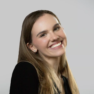 Portrait photo of a young woman with long light hair, wearing black clothing, smiling broadly and looking at the camera against a light background. She is wearing heart-shaped earrings.