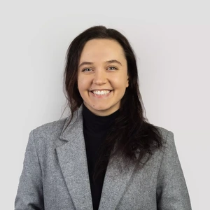 Portrait photo of a young woman with dark hair, wearing a grey blazer and a black turtleneck, smiling broadly at the camera against a light background.