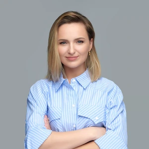 A woman with shoulder-length blonde hair, wearing a light blue striped shirt, stands with arms crossed and looks at the camera with a soft smile against a grey background.