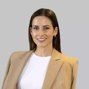 Portrait photo of a woman with long dark hair, wearing a beige pinstripe blazer and a white top. She is smiling and wearing gold earrings. The background is grey.