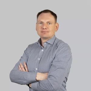 Portrait photo of a man with short dark hair, wearing a white shirt with fine blue stripes. He is standing with arms crossed and looking at the camera with a confident, serious expression. The background is neutral light grey.