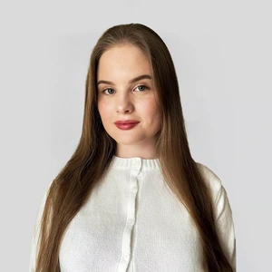 A young woman with long brown hair, wearing a white buttoned sweater, looks at the camera with a calm, neutral expression against a light background.