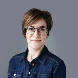 Portrait photo of a woman with short brown hair, wearing round black-rimmed glasses and a denim shirt-style jacket. She is gently smiling. The background is neutral dark grey.