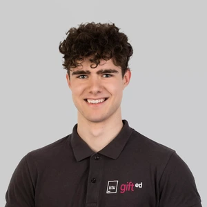 A young man with curly dark hair, wearing a dark polo shirt with the “ktu gifted” logo, is smiling and looking at the camera against a light background.