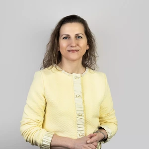 Portrait photo of a woman with medium-length wavy hair, wearing a light yellow jacket with decorative buttons. She is smiling gently, against a neutral light grey background.