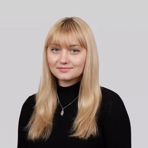 Portrait photo of a young woman with straight blonde hair and bangs, wearing a black high-neck sweater. She has a gentle smile and is wearing a pendant necklace, against a neutral grey background.