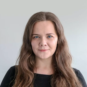 Portrait photo of a woman with long light brown hair, wearing a black top. She is looking directly at the camera with a calm expression, against a light neutral background.