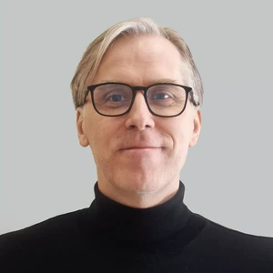 Portrait photo of a middle-aged man with light hair and glasses, wearing a black turtleneck, looking at the camera with a slight smile against a light background.