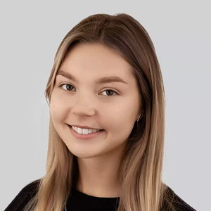 Portrait photo of a young woman with light hair, wearing a black top. She is smiling and wearing small earrings. The background is grey.