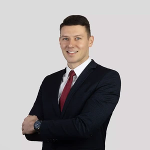 Portrait photo of a young man with short hair, wearing a dark suit, white shirt, and a red tie, smiling at the camera with arms crossed against a light background. He is wearing a watch on his wrist.