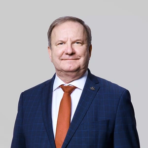 Portrait photo of an older man with light hair, wearing a dark blue checkered suit, white shirt, and an orange tie. The background is neutral gray. A small pin is attached to the lapel of his jacket.