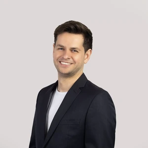 Portrait photo of a man with short dark hair, wearing a dark blazer and a white T-shirt. He is smiling at the camera, with a light neutral background.