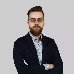 Portrait photo of a young man with a beard and glasses, short hair neatly styled. He is wearing a dark blazer and a white shirt with a small patterned design. Standing with arms crossed and looking at the camera with a serious expression. The background is neutral light grey.