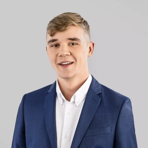 A young man with short blonde hair, wearing a blue suit and white shirt, is smiling and looking at the camera against a light background.