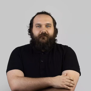 Portrait photo of a man with a full beard and curly hair, wearing a black polo shirt, looking directly at the camera with arms crossed against a light background.
