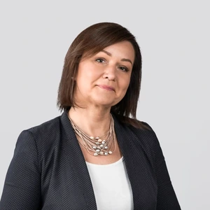 Portrait photo of a middle-aged woman with straight, shoulder-length dark brown hair. She is wearing a dark, subtly patterned blazer, a white top and a multi-strand necklace with pearls. She looks into the camera with a soft, calm expression. The background is light gray and neutral.
