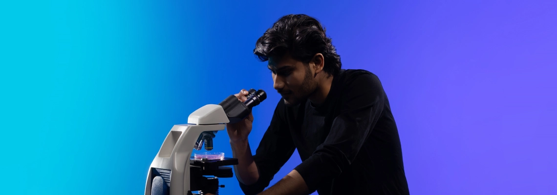 A young man dressed in black is looking through a microscope, focusing on samples placed on the stage. The background transitions from bright cyan to deep blue and violet. No visible text on the image.