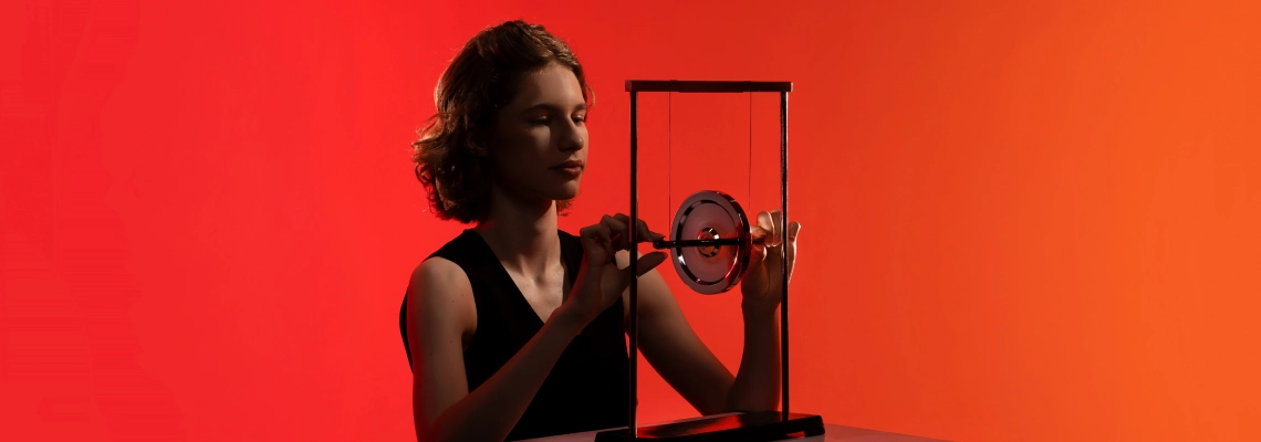 A young woman dressed in black is sitting at a table and adjusting a scientific apparatus with a suspended rotating disc. The background transitions from red to orange. No visible text on the image.