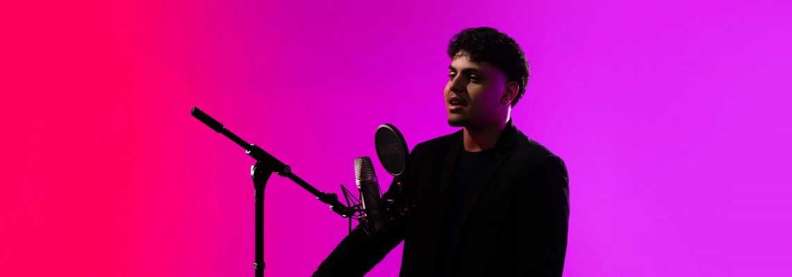 A young man dressed in black is standing in front of a professional microphone setup, as if speaking or recording. The background transitions from bright pink to purple. No visible text on the image.