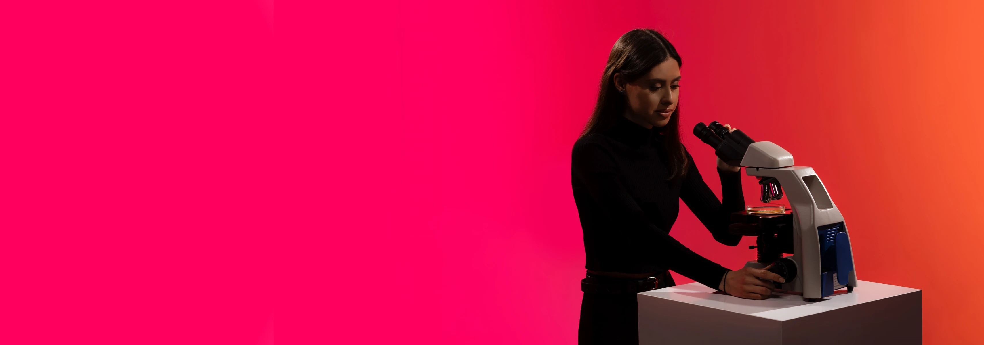A young woman stands against a pink background observing a sample through a microscope suitable for chemical technology research.
