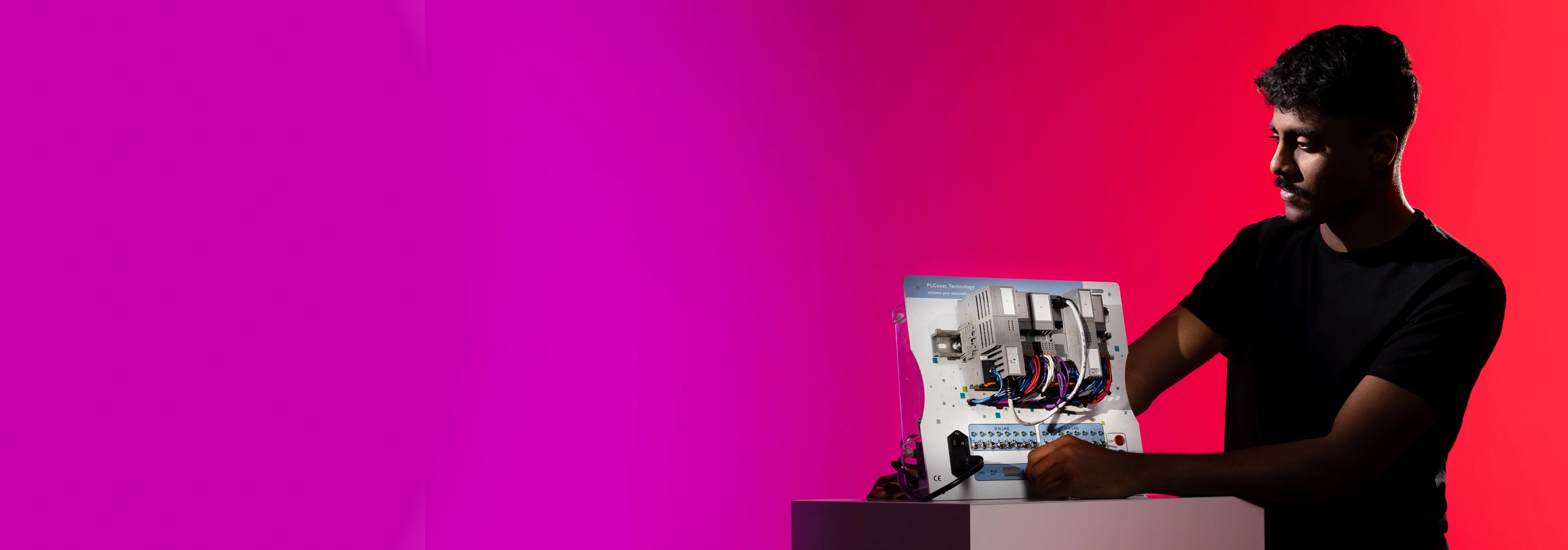 A male student stands against a pink background working with an automation control module suitable for electrical and electronics research.