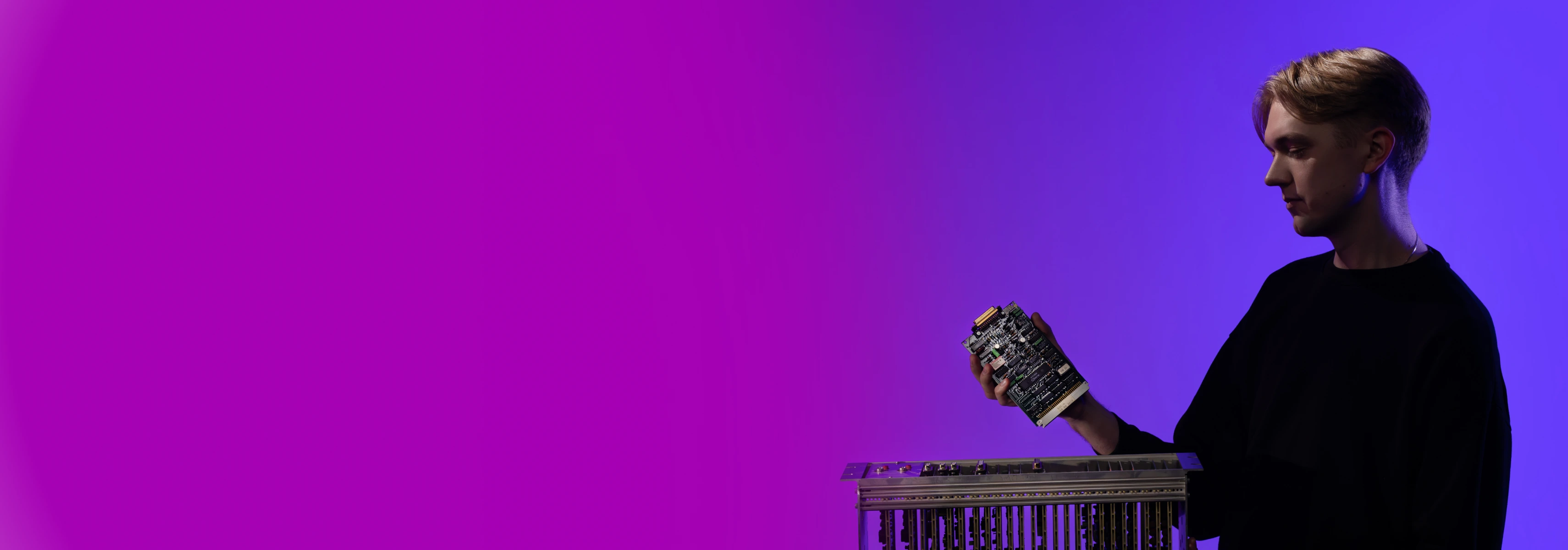 A male student stands against a violet background holding an electronic circuit board suitable for the activities of the Faculty of Informatics.