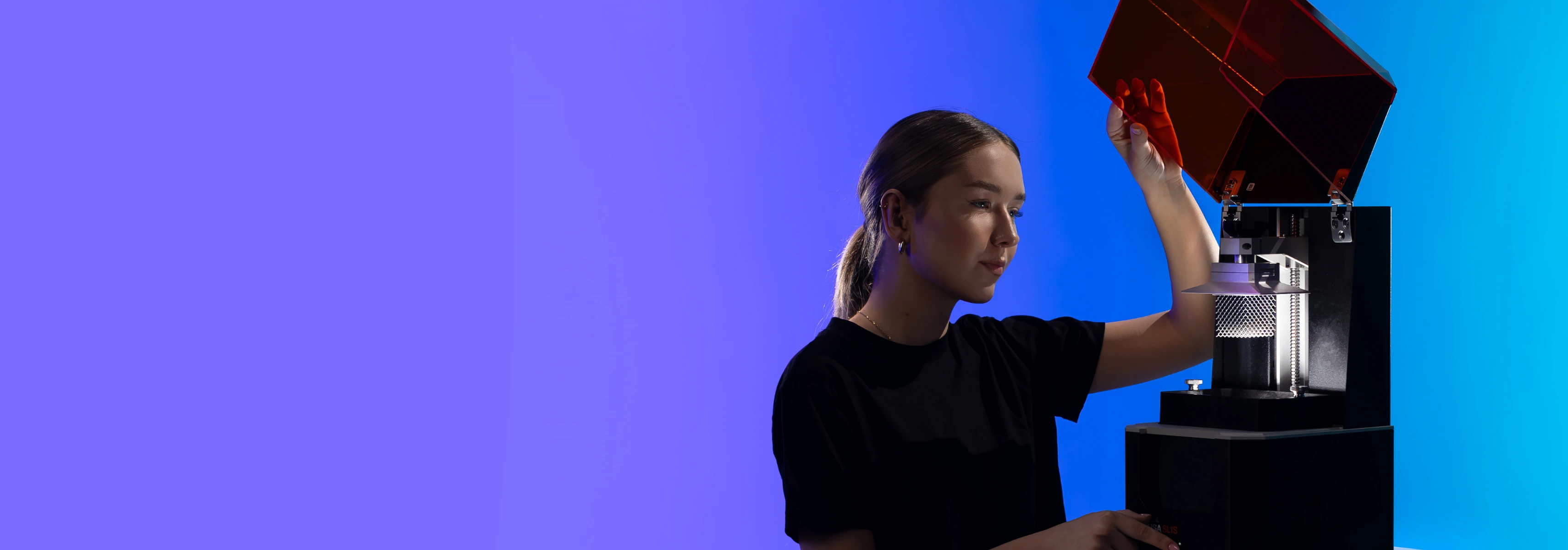 A young woman stands against a blue background observing a working 3D printer suitable for mechanical engineering and design activities.