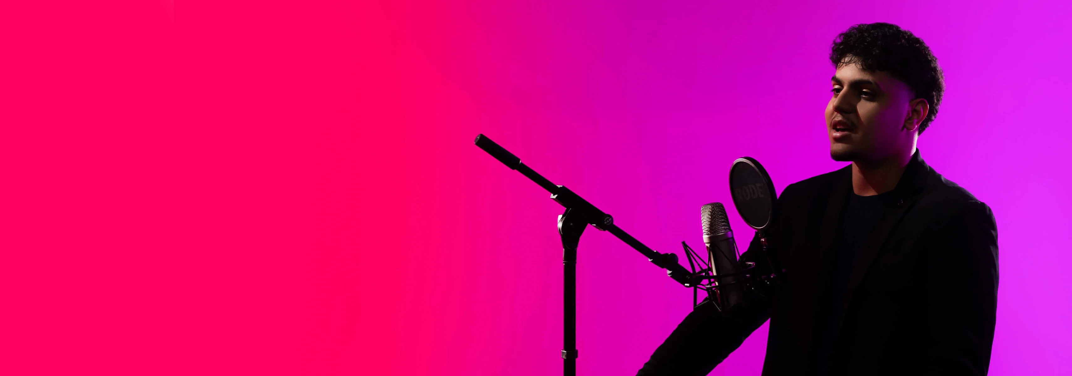 A young man stands against a violet background speaking into a microphone on a stand suitable for activities in the fields of social sciences, arts or humanities.