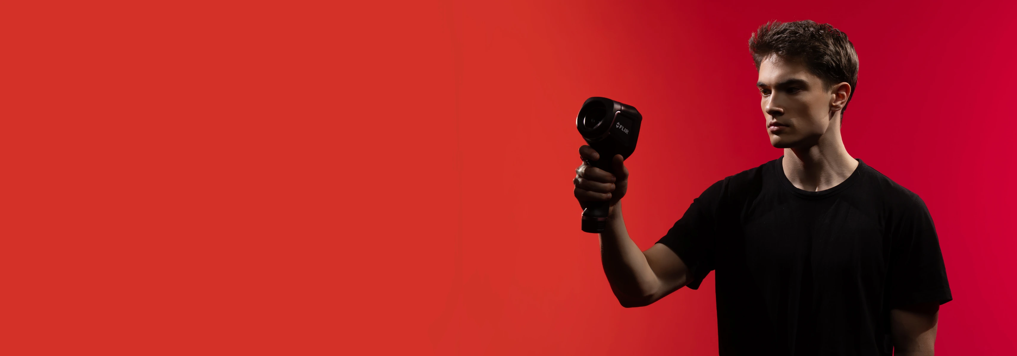 A young man stands against a red background using a thermal imaging camera suitable for environmental engineering research.