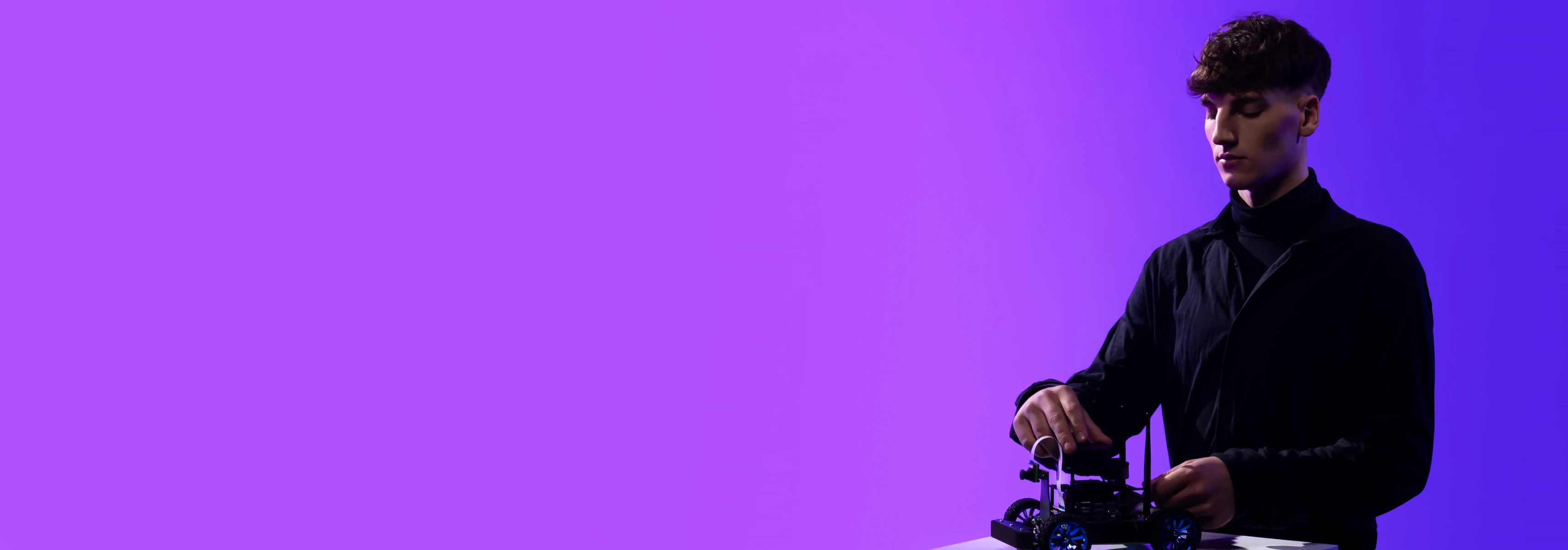 A young man stands against a violet background operating a small mobile robot suitable for studies combining technology and business.