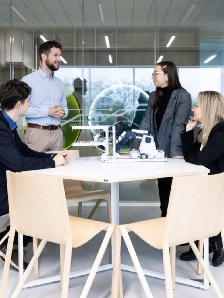 Group of PhD students standing around white table in bright, modern space collaborating and discussing research – doctoral studies collaboration culture, scholarships and funding opportunities at Kaunas University of Technology.