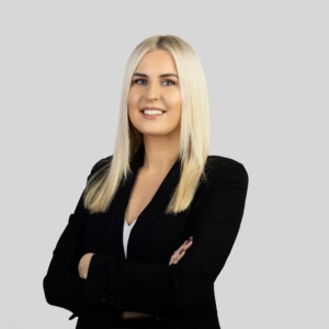 A young blonde woman with straight, shoulder-length hair, wearing a black blazer and a light-colored blouse, standing with her arms crossed and a professional smile, photographed against a neutral background.