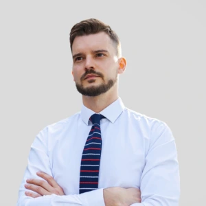 A young man with short dark hair, a mustache and beard, wearing a light shirt with a dark tie, arms crossed in front, smiling professionally, photographed against a neutral background.