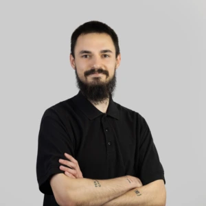 A young man with short dark hair, a mustache and beard, wearing a dark polo shirt, arms crossed in front, smiling professionally, photographed against a neutral background.