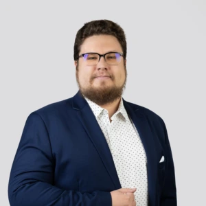 A young man with short dark hair, wearing glasses and a beard, dressed in a dark blue blazer and a light patterned shirt, with a calm and professional expression, photographed against a neutral background.