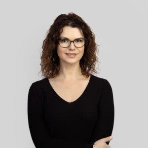 A young woman with shoulder-length wavy brown hair, wearing dark glasses, a black dress, arms crossed in front, smiling professionally, photographed against a neutral background.