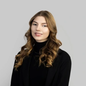 A young woman with long, wavy, shoulder-length brown hair, wearing a black blazer and black blouse, smiling professionally, photographed against a neutral background.