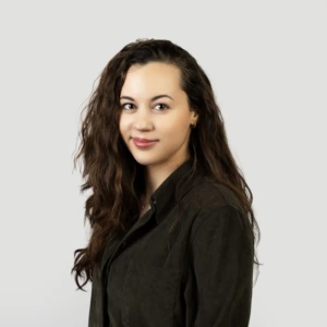 A young woman with long, wavy, shoulder-length dark hair, wearing a dark blazer, smiling professionally, photographed against a neutral background.
