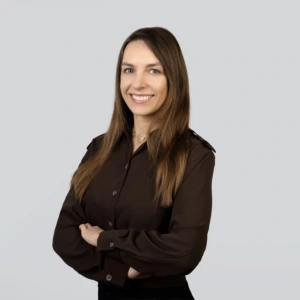 A young woman with long, straight brown hair, wearing a dark brown blouse, standing with her arms crossed and a professional smile, photographed against a neutral background.