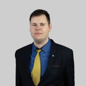 A young man with short dark hair, wearing a black blazer, a blue shirt, and a yellow tie, with a calm and professional expression, photographed against a neutral background.